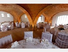 Ein rustikaler Veranstaltungsraum mit freiliegenden roten Backstein-Gewölbedecken, großen Fenstern und gedeckten Tischen mit weißen Stuhlhussen und kunstvoll gefalteten Servietten. Der Raum ist für eine festliche Veranstaltung vorbereitet.