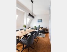 Modernes Wohnzimmer mit Essbereich, einem Holztisch und gepolsterten Stühlen auf Holzfußboden. Im Hintergrund ein graues Sofa, dekorative Kissen, eine Stehleuchte und ein kleiner Teppich; an der Decke eine Hängelampe mit mehreren Glaskugeln.