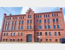 Backstein-Gebäude mit vier Stockwerken und neogotischen Elementen, einschließlich Spitzbögen und dekorativer Ziegelornamentik. Vor dem Gebäude befinden sich ein Basketballkorb, ein Kiesboden und Lichterketten, die im Vordergrund aufgehängt sind.