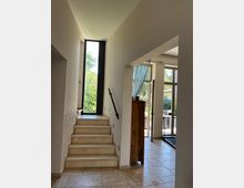 Heller Innenraum mit einer Treppe aus beigem Stein, die zu einem großen vertikalen Fenster mit Blick auf Bäume führt. Rechts befindet sich ein Raum mit Glasfenstern und Türen, die nach außen führen, sowie ein dunkler Vorhang, der teilweise das Fenster verdeckt.