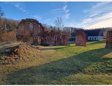 Ruine eines Backsteingebäudes mit großen, bogenförmigen Fenstern auf einer Grasfläche, umgeben von Bäumen; im Hintergrund ein Fachwerkhaus.
