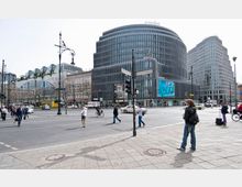 Straßenecke am Kurfürstendamm in Berlin, mit einem modernen Gebäudekomplex im Hintergrund, einschließlich des Swissôtel und einer Karstadt-Filiale. Mehrere Personen überqueren eine breite Kreuzung, umgeben von Autos, Fahrrädern und historischen Straßenlaternen.
