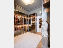 Eleganter begehbarer Kleiderschrank mit großen, verglasten Schränken, die beleuchtete Regale mit Kleidung und Schuhen zeigen. Im Vordergrund steht eine Schaufensterpuppe in einem verzierten Kleid, und die Decke ist mit dekorativem Stuck versehen.