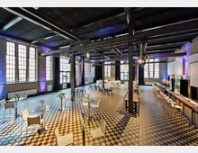 Großer Veranstaltungssaal mit industriellem Stil, gekennzeichnet durch ein Schachbrettmuster am Boden und große Fenster mit Metallrahmen. Es gibt vereinzelte Tische und Stühle, eine Bar sowie moderne Lichtinstallationen an den schwarzen Stahlträgern der Decke.