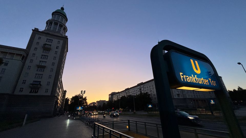 Blick in der Dämmerung auf eine breite Stadtstraße mit mehreren Fahrspuren und angrenzenden Wohn- und Geschäftsbauten. Im Vordergrund steht ein beleuchtetes U-Bahn-Schild „U Frankfurter Tor“, links ragt ein hohes Gebäude mit Turmkuppel in den Himmel.