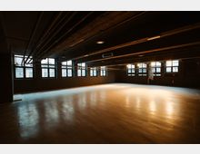 Ein leerer, dunkler Raum mit Betonboden, großen Fenstern an einer Wand und freiliegenden Rohren an der Decke. Die Beleuchtung ist gedämpft, und einige Fenster lassen Tageslicht herein.