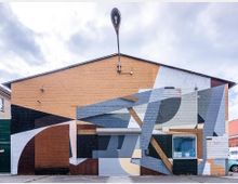 Backstein-Gebäude mit einer großen abstrakten Wandmalerei, die geometrische Formen in Beige-, Grau-, Braun- und Schwarztönen zeigt; das Gebäude hat ein Schrägdach, ein Rolltor und ein kleines Fenster auf der rechten Seite. Auf dem Dach ist eine skulpturale Straßenlaterne angebracht.