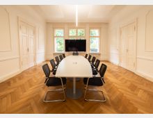 Heller Besprechungsraum mit einem langen weißen Konferenztisch, umgeben von schwarzen Stühlen. Der Raum hat einen hölzernen Fischgrätboden, große Fenster mit Blick nach draußen und eine Wand mit einem montierten Flachbildschirm.