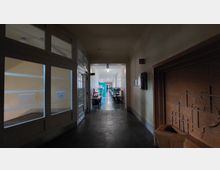 Ein Flur in einem Gebäude mit seitlich verglasten Türen auf der linken Seite, die zu einem beleuchteten Raum führen, und einer verzierten Wandnische aus Stein auf der rechten Seite. Im Hintergrund sind Büromöbel in einem helleren Bereich sichtbar.