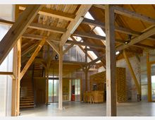 Innenraum eines renovierten Scheunengebäudes mit sichtbaren Holzbalken und einem hohen, offenen Dachstuhl. Im Raum befinden sich eine Holztreppe, eine Bar aus Holzlatten und Wände aus einer Kombination von Sichtziegeln und transluzenten Materialien.