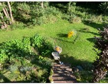 Garten mit gepflegtem Rasen, Blumenbeeten und Bäumen am Rand. Im Vordergrund befinden sich Holztreppen, zwei Stühle und ein Tisch, die im Schatten stehen.