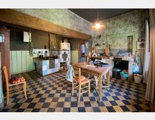 Rustikale Küche mit kariertem Boden in Schwarz und Beige, einem hölzernen Esstisch mit Stühlen und einer Küchenzeile mit Kacheln in Blau und Weiß. An der Wand hängen Werkzeuge und Pflanzen, und ein großer Holzbalken verläuft quer durch den Raum.