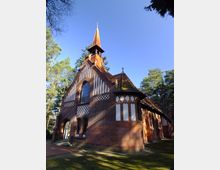 Rotes Backsteingebäude einer historischen Kirche mit kleinen Fenstern und weiß verputzten Fachwerkelementen an der Fassade. Im Hintergrund stehen hohe Bäume bei klarem, sonnigem Himmel.