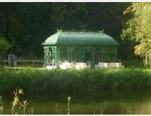 Grüne, filigrane Metall-Pavillon-Struktur mit gläsernem Dach, umgeben von Bäumen und weichem Gras. Im Vordergrund ein Teich und Schilf, während weiße Sitzmöbel im Pavillonbereich stehen.