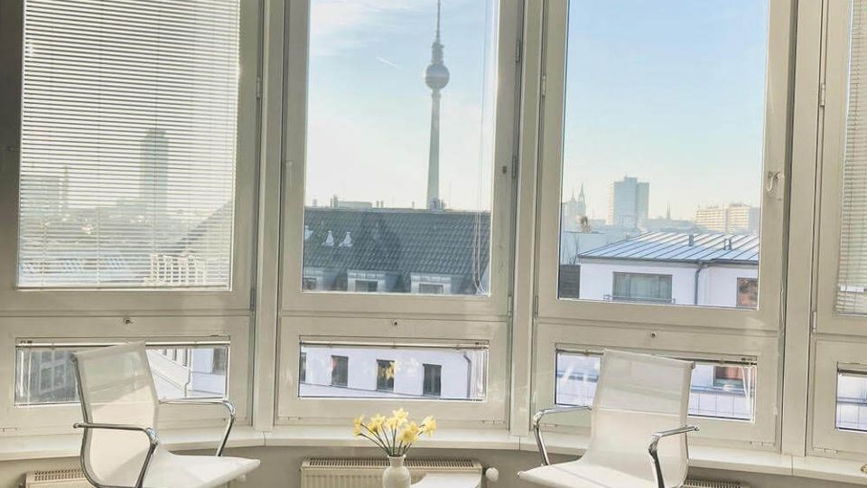 Bright modern office lounge with curved floor-to-ceiling corner windows fitted with blinds, overlooking a city skyline with a tall TV tower. Two white swivel chairs and small curved side tables sit on dark wood flooring beside low radiators under the windows.