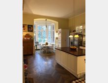 Große, helle Küche mit cremefarbenen Wänden und Holzfußboden, ausgestattet mit einer zentralen Kücheninsel und Hängeleuchten. Im Hintergrund befinden sich ein Erker mit hohen Fenstern und ein runder Esstisch mit weißem Stuhlset.