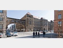 Straßenszene mit einem großen historischen Steinbauwerk in Berlin, das durch ein steinernes, mit Fenstern versehenes Bogenelement zwei Gebäude verbindet. Vor dem Gebäude sind Menschen, Autos und Verkehrsschilder zu sehen.