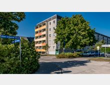 Mehrstöckiges Wohngebäude mit farbigen Balkonen im Vordergrund, umgeben von Bäumen und parkenden Autos. Straßenschilder weisen auf die Kreuzung der Albert-Schweitzer-Straße und Robert-Koch-Straße hin; eine "Tempo 30"-Zone ist ebenfalls zu sehen.