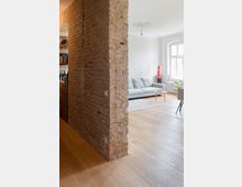 Wohnraum mit modernem Design, unterteilt von einer freistehenden Wand aus alten Backsteinen. Der Raum verfügt über Holzböden, eine graue Couch, ein großes Fenster mit weißem Rahmen und minimalistische Einrichtung.