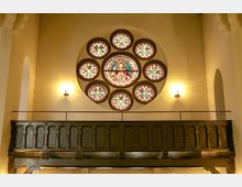 Innenraum einer Kirche mit einer großen, runden Rosettenfenstergruppe aus buntem Glas, die zentral im oberen Bereich der Wand eingesetzt ist. Darunter ein dunkles hölzernes Geländer einer Empore, seitlich flankiert von zwei Wandlampen, die den Raum beleuchten.