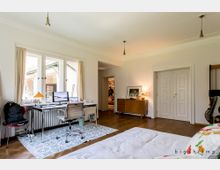 Helles Schlafzimmer mit Arbeitsbereich, ausgestattet mit einem Schreibtisch, Bürostuhl und zwei Monitoren vor großen Fenstern mit Vorhängen. Im Raum befinden sich außerdem eine weiße Doppeltür, eine Kommode und Parkettboden.