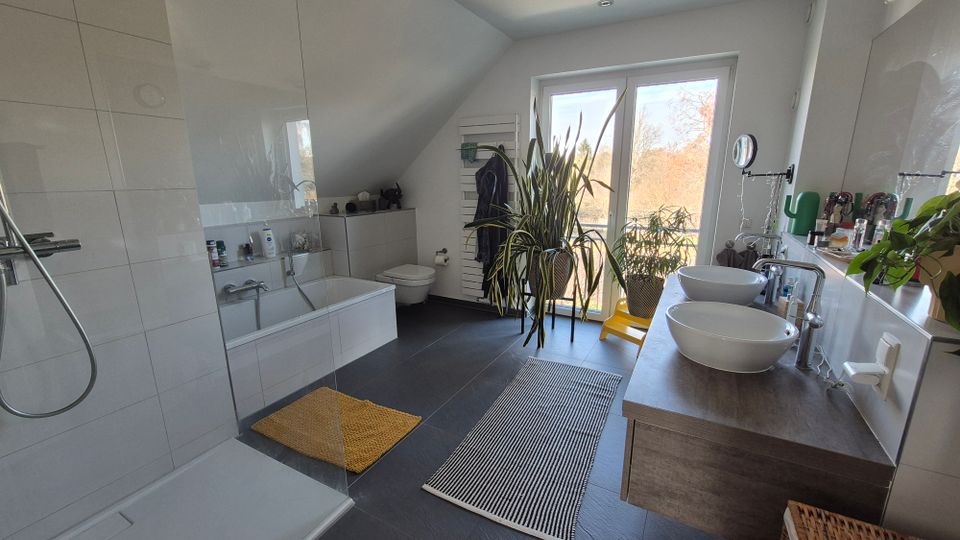 Modernes Badezimmer mit bodengleicher Dusche, weißer Badewanne und Doppelwaschbecken auf einem Holzunterschrank. Große Fenster mit Blick ins Freie lassen viel Tageslicht herein, ergänzt durch Zimmerpflanzen und dunkle Bodenfliesen.