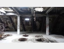 Ein leerer, verlassener Raum mit einer schrägen Betonwand, teilweise eingestürztem Dach und mehreren kreisförmigen Löchern im Boden. Graffiti bedecken die Betonstützen, und durch ein kleines Fenster fällt Licht in den ansonsten düsteren Raum.