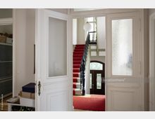 Blick in ein historisches Treppenhaus mit roten Teppichläufern, einer Holztreppe mit kunstvoll verziertem schwarzen Geländer und einem großen verglasten Eingangsbereich mit Holztüren. Die Umgebung ist durch weiße Türen mit Milchglaseinsatz eingerahmt.