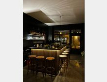 Eine moderne Bar mit einer Theke aus vertikal angeordneten Holzlatten, umgeben von hohen Hockern mit runden Holzsitzen. Im Hintergrund befinden sich Regale mit Flaschen und Gläsern, sowie eine große Fensterfront, die nach draußen blickt.