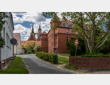 Ein Krankenhausgebäude aus rotem Backstein mit weißen Fenstern, umgeben von Bäumen und gepflegter Grünfläche. Im Hintergrund sind die Türme einer historischen Kirche zu sehen, daneben ein Schild mit der Aufschrift „Johanniter-Krankenhaus im Fläming“.