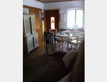 Wohnzimmer mit ländlichem Stil, das einen runden Esstisch mit hellen Stühlen zeigt. Im Raum befinden sich ein rustikaler Holzschrank, eine Couch, sowie ein Fenster und eine Tür mit farbigen Glaselementen.