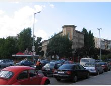 Straßenszene mit mehrstöckigem historisch wirkendem Gebäude im Hintergrund, Bäumen und einem kleinen Kiosk mit roten Dächern. Im Vordergrund dichter Autoverkehr und ein roter VW Käfer auf der Straße.