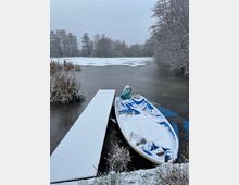 Ein zugefrorener See mit einem schneebedeckten Steg und einem kleinen Ruderboot im Vordergrund. Umgeben von schneebedeckten Bäumen und Schilf am Ufer.