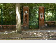 Rote Backsteinpfeiler mit einer grünen Metallpforte und seitlicher Zaunanlage, umgeben von dichtem Bewuchs. Im Vordergrund ein Baumstamm und ein schmaler Gehweg mit Laub auf dem Boden.