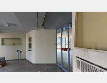 Ein leerer Innenraum mit beige gestrichenen Wänden, einer Glastür mit rotem Quergriff, und großen Fenstern im Hintergrund, die Blick auf die Außenwelt bieten. Decke zeigt Abnutzungserscheinungen, und auf dem Boden liegen vereinzelt Gegenstände wie Holzpaletten.