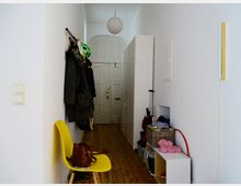 Schmaler Flur mit Holzboden, weiß gestrichenen Wänden und einer hohen Decke. Links an der Wand befindet sich eine Garderobe mit Jacken und einem Fahrradhelm, darunter ein gelber Stuhl mit einer Ledertasche. Rechts stehen ein Schrank, eine kleine Regalbox mit einem Korb, Haushaltsgegenstände und eine Tür am Ende des Flurs.