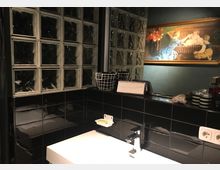 Ein modernes Badezimmer mit schwarzem Fliesenspiegel und Waschbecken. Im Hintergrund sind Glasbausteine, ein Wandbild mit einem klassischen Gemälde sowie ein Regal mit Dekoartikeln und Haushaltsgegenständen zu sehen.