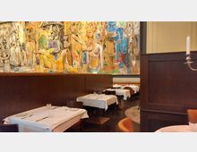 Restaurant-Innenraum mit gedeckten Tischen, weißer Tischwäsche und elegantem Holzmobiliar. An der Wand ein großes, buntes Wandgemälde mit abstrakten Figuren.