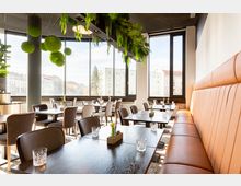 Ein modernes Restaurant mit großen Fenstern, die Blick auf umliegende Gebäude bieten. Die Einrichtung umfasst dunkle Holztische, beige und braune Stühle sowie eine lange gepolsterte Sitzbank; Dekorationen wie Glasvasen und hängende Grünpflanzen zieren den Raum.