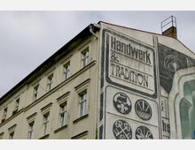 Außenansicht eines mehrstöckigen, beige gestrichenen Gebäudes mit einer großflächigen Wandbemalung, die den Schriftzug „Handwerk & Tradition“ sowie Symbole für handwerkliche Tätigkeiten zeigt. Die Fassade ist leicht verwittert, und der Himmel ist bewölkt.