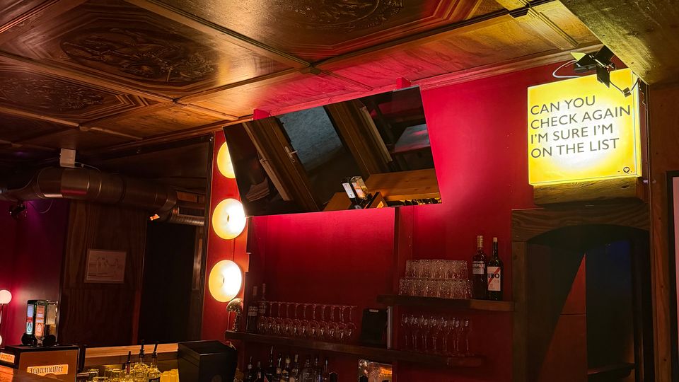 Innenraum einer Bar mit langem Holzthekenbereich, roten Wänden und Regalen mit Flaschen und Gläsern. An der Decke sind dekorative Holzpaneele zu sehen, daneben Spiegel, runde Wandleuchten und ein beleuchtetes Schild mit Text.