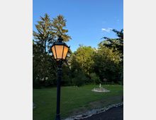 Ein Park bei Sonnenuntergang mit einer beleuchteten Laterne im Vordergrund, einem Rasenbereich, mehreren Bäumen im Hintergrund und einem kleinen Brunnen mit einer Skulptur. Der Himmel ist klar und blau.