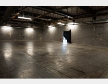 Großer, leerer Industriehallenraum mit grauen Betonblockwänden und poliertem Betonboden. Offene Decke mit sichtbaren Trägern, Kabeln und Lüftungsrohren sowie hängenden Leuchtstoffröhren; seitlich eine Türöffnung mit schwarzem Vorhang.