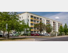 Mehrstöckiges Wohngebäude mit mehreren Balkonen und einer überwiegend hellen Fassade mit gelben Akzenten. Im Vordergrund sind geparkte Autos, Gehwege, Bäume und Straßenschilder zu sehen.