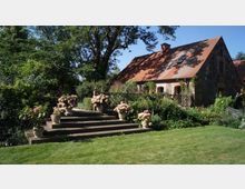 Steingebäude mit roten Ziegeldächern in ländlicher Umgebung, umgeben von einem gepflegten Garten mit Blumenbeeten, Rasenflächen und einer breiten Steintreppe, die mit Topfpflanzen dekoriert ist. Im Hintergrund sind Bäume und Sträucher zu sehen.