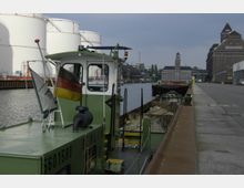 Ein industrieller Hafen mit einem grünen Arbeitsschiff im Vordergrund, welches mit der deutschen Flagge versehen ist. Im Hintergrund sind weiße Tanks, Lagergebäude aus Backstein und ruhiges Wasser zu sehen.