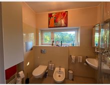 Modernes Badezimmer mit beigefarbenen Fliesen, wandhängendem WC und Bidet sowie Waschbecken neben einer Glasduschkabine. Ein breites Fenster mit Blick ins Grüne und ein abstraktes Bild darüber prägen den Raum.