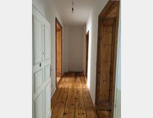 Ein schmaler Flur in einem Wohnraum mit Holzdielenboden und weißen Wänden. Rechts und links befinden sich Türen mit hölzernen Rahmen, und am Ende des Flurs ist eine Wand zu sehen.