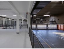 Parkdeck in einer Tiefgarage mit Betonböden, weißen Wänden und Metallgeländern. Im Hintergrund sind große Rolltore mit Verglasung sowie helle Beleuchtung durch Tageslicht erkennbar.