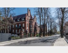 Backsteingebäude im neugotischen Stil mit spitzen Giebeln, großen Fenstern und dekorativen Fassadenelementen, entlang einer von Bäumen gesäumten Straße. Im Vordergrund ein Gehweg, ein Zaun und parkende Autos, im Hintergrund einige Wohnhäuser.